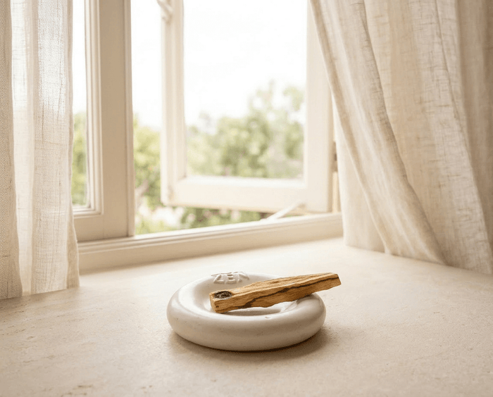 Palo Santo Stick liegt auf einer ZoaZen Keramikschale auf der Fensterbank – offenes Fenster mit Leinenvorhängen und Blick ins Grüne