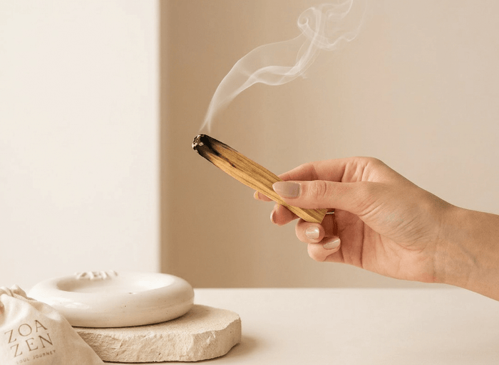 Hand hält einen glimmenden Palo Santo Stick mit aufsteigendem Rauch – im Hintergrund ZoaZen Keramikschale und Baumwollbeutel